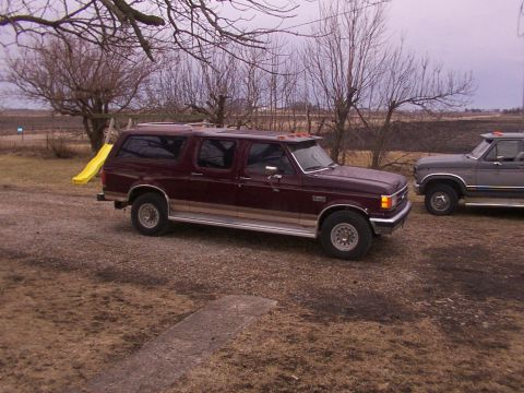 1988 Ford C-350 (Centurion Conversions) - Two Ford trucks are parked in a gravel lot with bare trees and fields in the background.