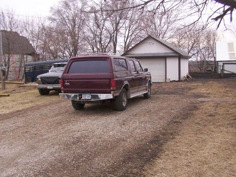 1988 Ford C-350 (Centurion Conversions) - Maroon Ford Bronco Centurion with aftermarket bumper and roof rack is parked in a gravel lot near a garage.