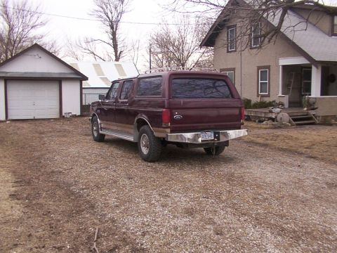 1988 Ford C-350 (Centurion Conversions) - Maroon Ford Bronco SUV with camper shell parked in a gravel driveway next to a house and garage.