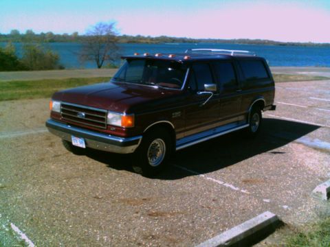 1988 Ford C-350 (Centurion Conversions) - A maroon Ford F-Series SUV conversion is parked in a lot next to a body of water under a clear sky.