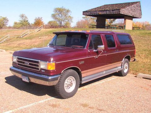 1988 Ford C-350 (Centurion Conversions) - A maroon SUV with tan trim is parked in a lot near a picnic shelter and stairs.