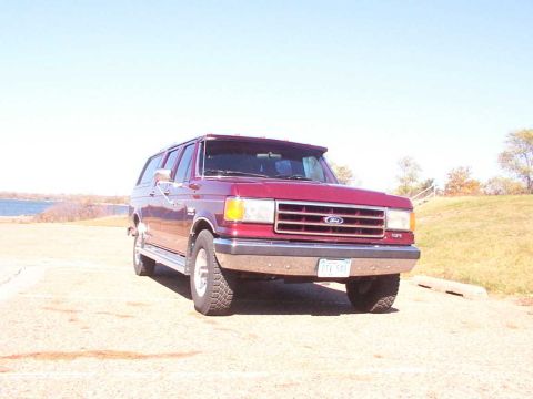 1988 Ford C-350 (Centurion Conversions) - A red SUV is parked on a dirt lot with a body of water and grassy hill in the background.