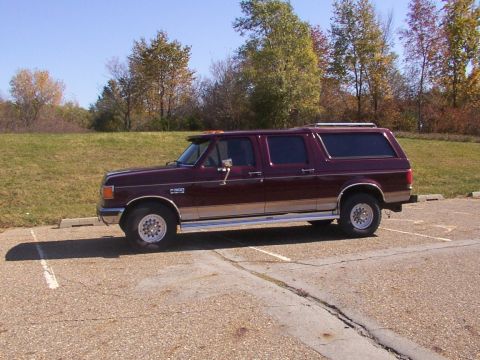 1988 Ford C-350 (Centurion Conversions) - A maroon Ford pickup truck with a camper shell is parked in a lot on a sunny day with trees in the background.