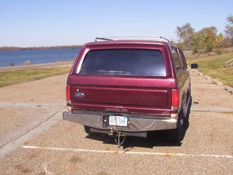 1988 Ford C-350 (Centurion Conversions) - A maroon Ford Bronco is parked near a body of water on a sunny day, showing its rear end and chrome bumper.