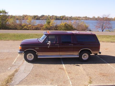 1988 Ford C-350 (Centurion Conversions) - A side view of a maroon Ford F-Series SUV conversion with tan trim, parked in a lot near a body of water.