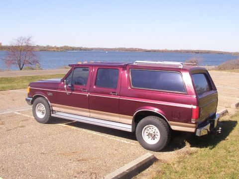 1988 Ford C-350 (Centurion Conversions) - Maroon extended cab pickup truck with a camper shell, parked in a lot near a body of water.