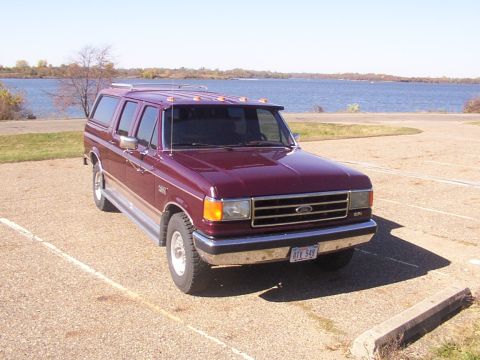 1988 Ford C-350 (Centurion Conversions) - Maroon Ford SUV conversion with camper shell parked near a body of water.