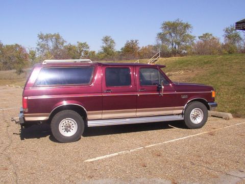 1988 Ford C-350 (Centurion Conversions) - Side view of a maroon SUV with tan trim and a roof rack parked in a gravel lot under a clear blue sky.