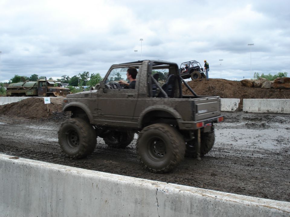 Muddy brown lifted Suzuki Samurai on off-road mud course, rear side view