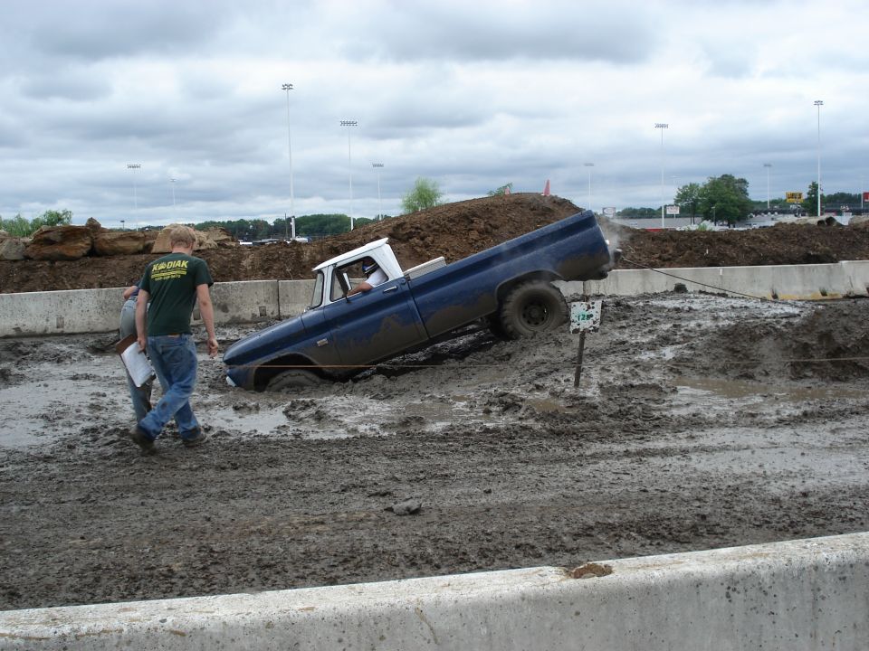 Blue pickup truck stuck in mud pit with two people standing nearby