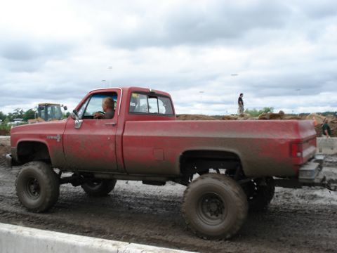 1988 Suzuki Samurai - A lifted red pickup truck navigates a muddy track under a cloudy sky.