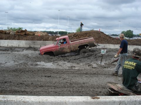 1988 Suzuki Samurai - A red pickup truck is stuck deep in mud during a mud bog event, with spectators nearby.
