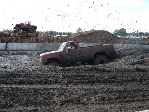 1988 Suzuki Samurai - A red pickup truck is deeply stuck in a mud bog, while another vehicle sits on a dirt embankment above.