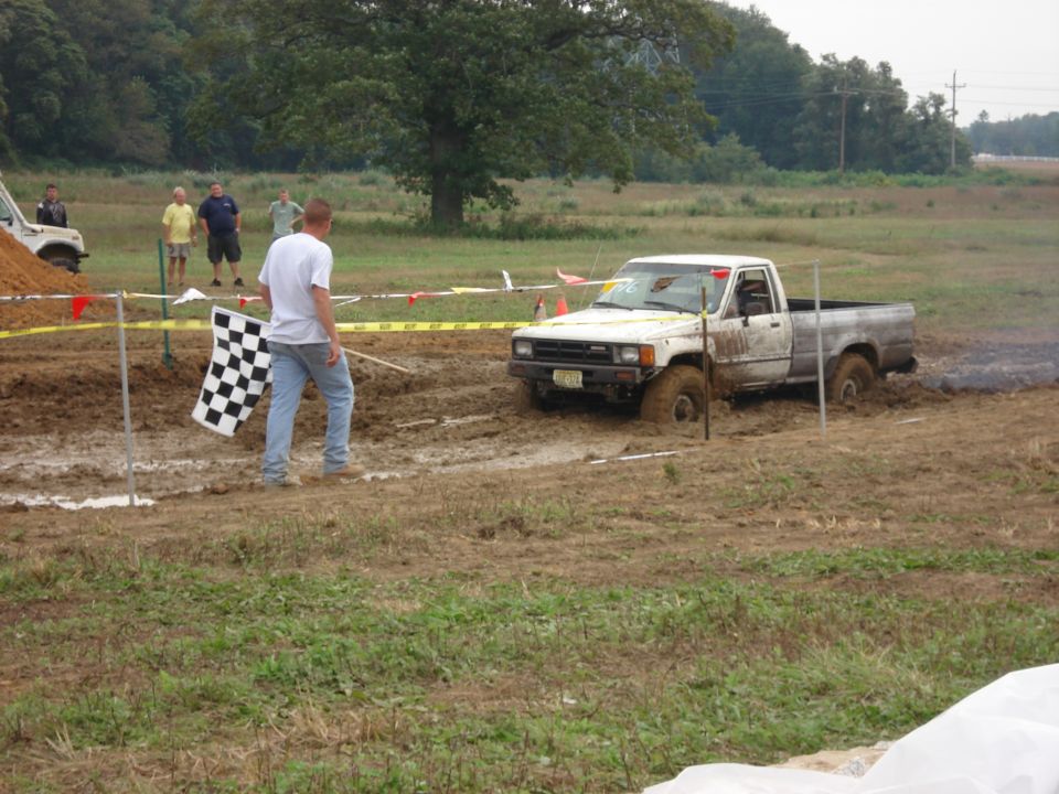 White pickup truck stuck in mud, with a person holding a checkered flag nearby. Another white SUV is visible in the background