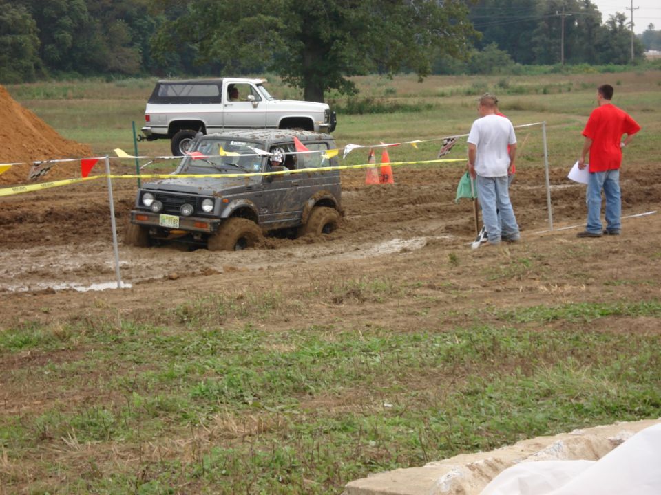 Dark gray 1988 Suzuki Samurai stuck in mud, front-left wheel submerged, with a white SUV in the background