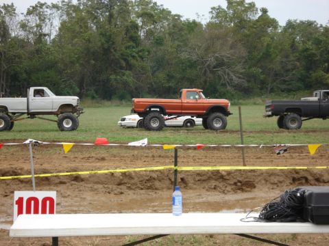 1988 Suzuki Samurai - Modified pickup trucks participate in a mud bogging event with a white car underneath one of the trucks.
