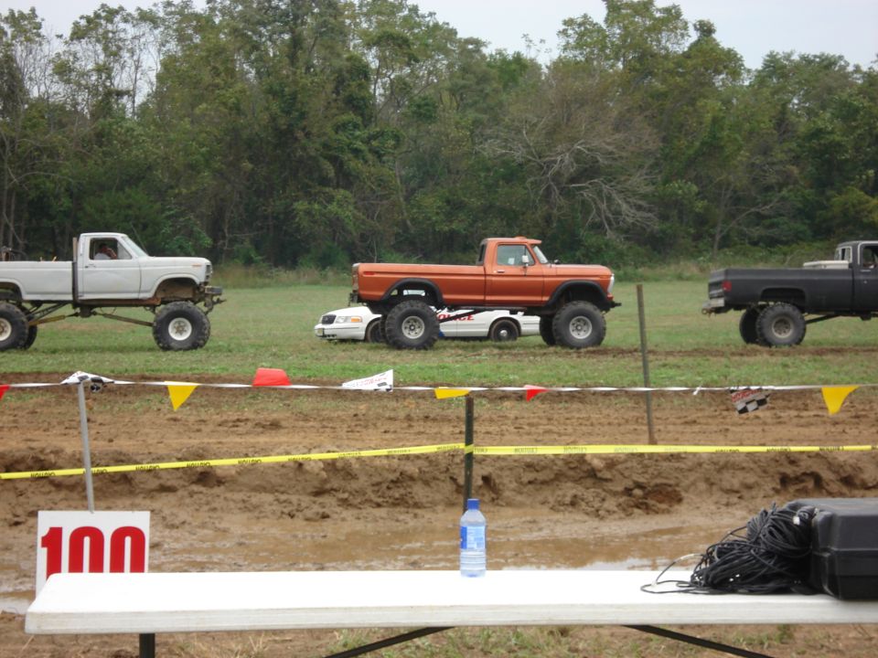 Three lifted pickup trucks in a mud bog, one orange truck is on top of a white car