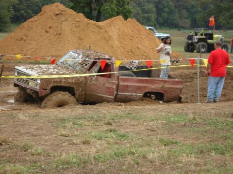 1988 Suzuki Samurai - A red pickup truck is stuck in a mud bog, with other vehicles and people in the background.