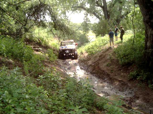 Light-colored 4x4 climbing a rutted muddy gully while three spectators watch