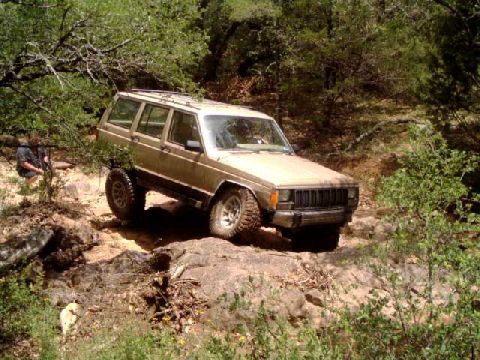 1984 Ford Bronco - A tan Jeep Cherokee XJ navigates a rough, rocky terrain surrounded by dense foliage.