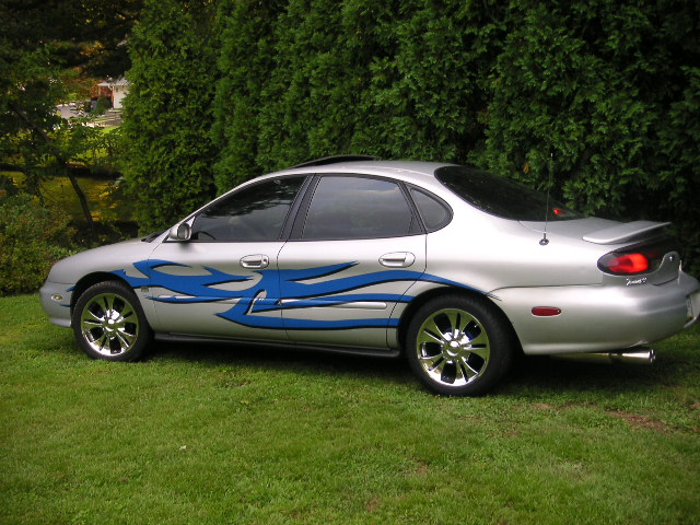 Silver sedan with blue flame decals on the side, parked on grass with trees in the background