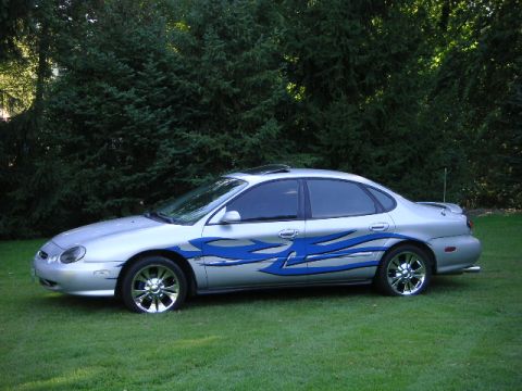 1999 Ford Taurus - A silver 1999 Ford Taurus sedan with custom blue flame decals and chrome wheels is parked on a grassy lawn in front of trees.