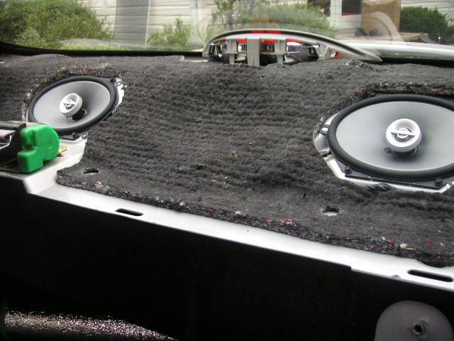 Car dashboard interior with exposed insulation and two oval speakers removed, showing wiring and a green tool