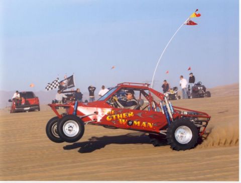 1993 Ford Taurus - Red flame-painted tube-chassis sand rail pulling the front wheels off the ground on a dune, lettered 'THE OTHER WOMAN' along the side panel…