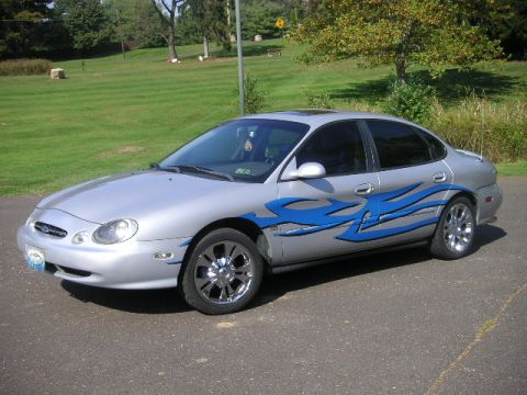 1999 Ford Taurus - A silver 1999 Ford Taurus sedan with custom blue flame decals and chrome wheels is shown from a three-quarter front angle.