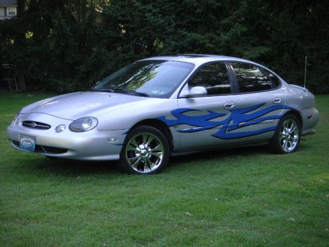 1999 Ford Taurus - A silver sedan with blue flame decals is parked on a grassy lawn with trees in the background.
