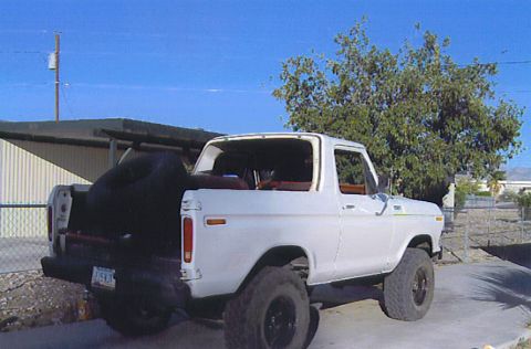 1990 Ford Bronco - white lifted Ford Bronco with its hardtop removed, parked beside a carport with a spare tire in the bed area.