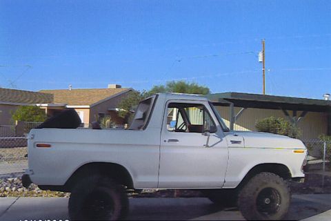 1990 Ford Bronco - white lifted Ford Bronco shown in side profile parked on a driveway with houses, a carport and clear blue sky behind.