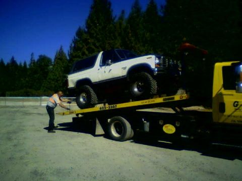 1986 Ford Bronco - A white Ford Bronco is loaded onto a yellow tow truck by a man.