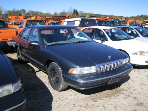 1991 Ford LTD Crown Victoria - A dark sedan is parked in a lot with other vehicles and orange trucks visible in the background.