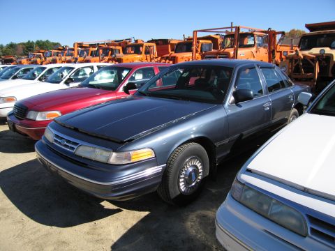 1991 Ford LTD Crown Victoria - A dark blue Ford Crown Victoria is parked in a lot, with its hood open, alongside other cars and large orange trucks.
