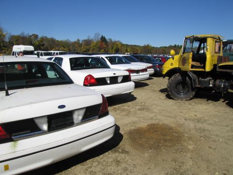 1991 Ford LTD Crown Victoria - Several white Ford Crown Victoria LTD sedans are parked in a lot, with a yellow truck visible on the right side of the frame.