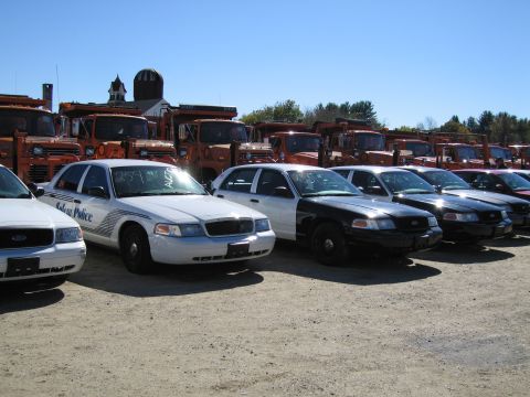 1991 Ford LTD Crown Victoria - Police cars are lined up in a lot, with a row of orange trucks behind them under a clear blue sky.