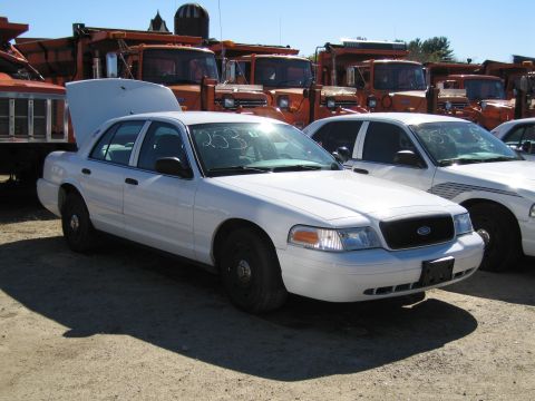 1991 Ford LTD Crown Victoria - A white sedan is parked in front of several large orange dump trucks, with another white sedan beside it.