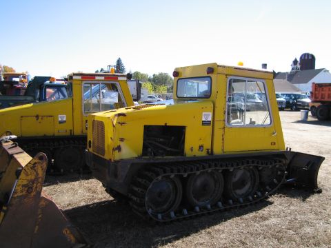 1991 Ford LTD Crown Victoria - Two yellow tracked vehicles are parked outdoors, one with a plow attachment in the foreground.