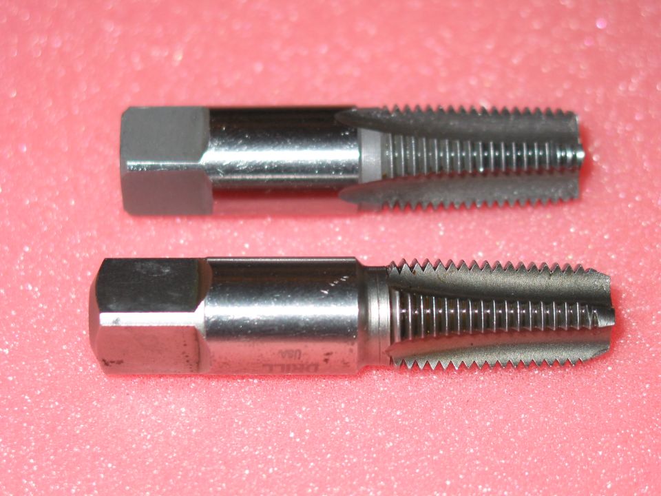 Two steel tapered pipe taps with square drive ends on a pink surface