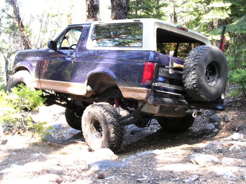 1994 Ford Bronco - A lifted Ford Bronco navigates a challenging, wooded trail with large tires and a spare mounted on the back.