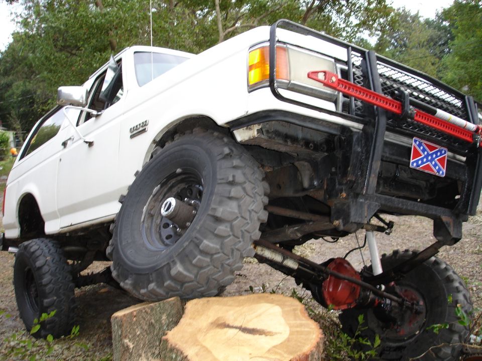 Lifted white Ford Bronco flexing over a tree stump on big mud tires with tube grille guard