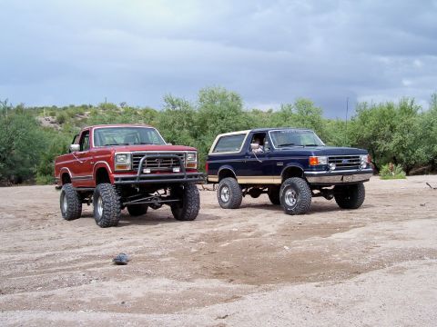 1991 Ford Bronco - Two lifted Ford trucks, a pickup and a Bronco, sit parked on a dirt lot under a cloudy sky.