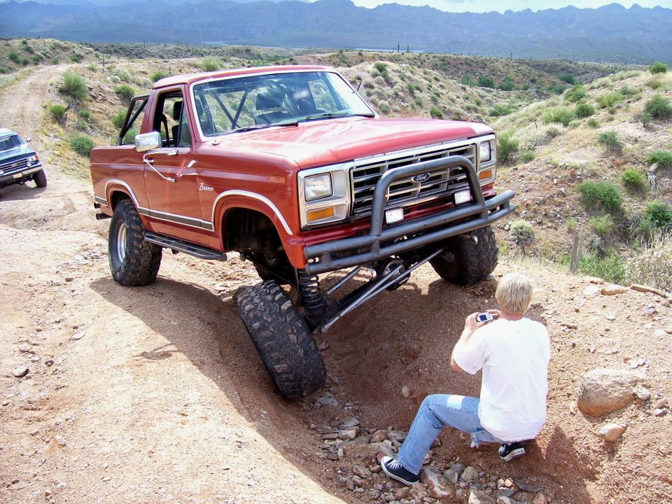 Red Ford Bronco with lifted suspension navigating rough terrain, person photographing