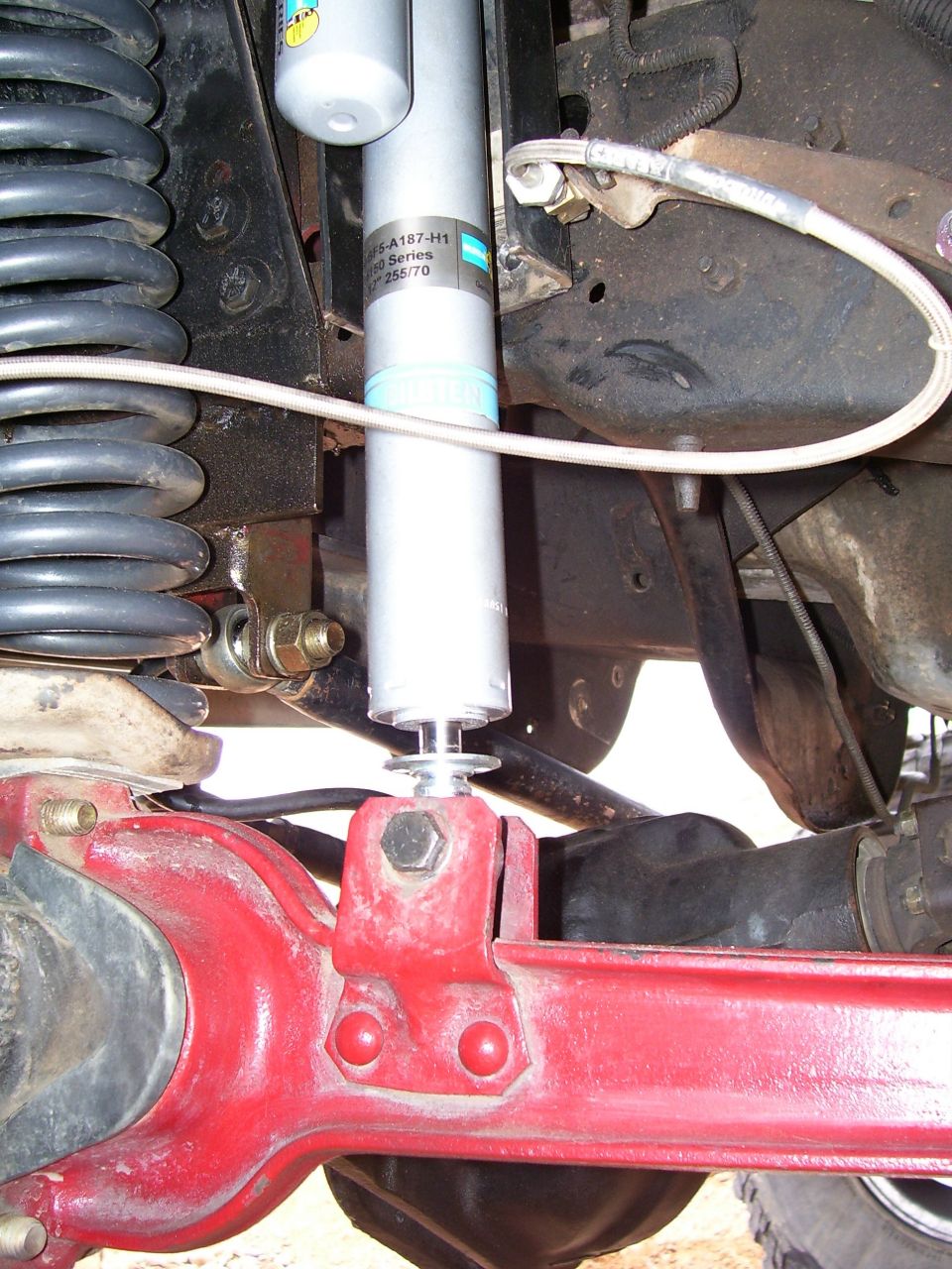 Close-up of a vehicle's rear suspension with a red axle, black coil spring, and a silver Bilstein shock absorber. Text on the shock reads 'Bilstein 5100 Series'