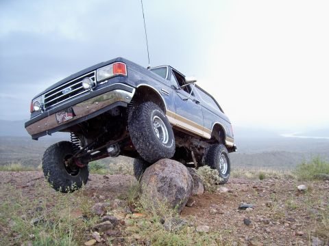 1991 Ford Bronco - A lifted blue Ford Bronco navigates rocky terrain under a cloudy sky.