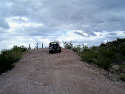 1991 Ford Bronco - A tan and black SUV navigates a dusty trail under a dramatic, cloudy sky, with cacti dotting the arid landscape.