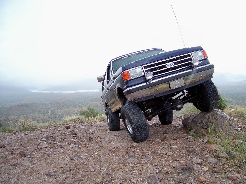 1991 Ford Bronco climbing a rock on a dirt hill with a lake in the background
