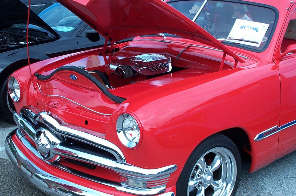 Red classic car with chrome grille and hood open, showing engine. Another black car is visible behind it