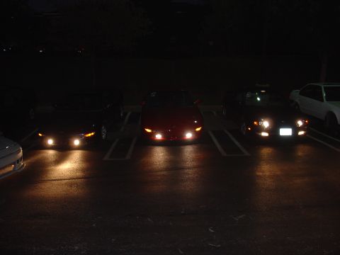1993 Ford Probe - Several sports cars are parked in a lot at night, their headlights illuminating the asphalt.